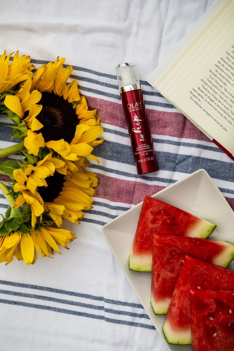 Olay Regenerist Whip review and summer self care routine featured by top D.C beauty blogger, Alicia Tenise | Self Care Routine by popular D.C. lifestyle blog, Alicia Tenise: image of a bottle of Olay Regenerist whip on a red, white, and blue stripe turkish towel next to a open book and a white plate with a watermelon slices on it.