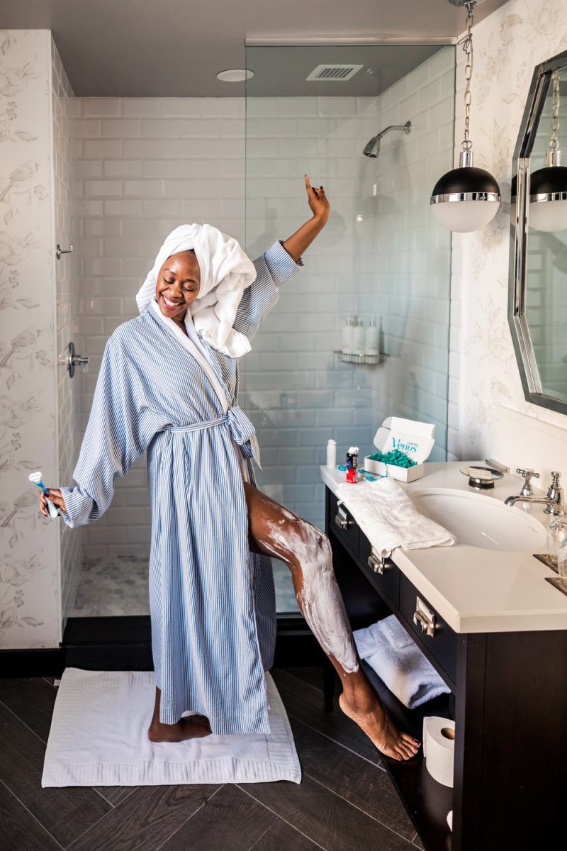 Self Care Ideas by popular DC lifestyle blogger, Alicia Tenise: image of a woman shaving her legs in her bathroom and wearing a blue and white striped bathrobe and a white towel wrap on her head.