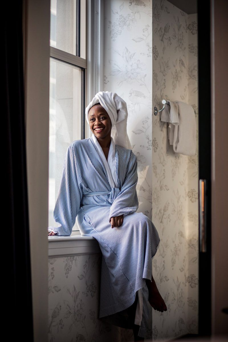 Self Care Ideas by popular DC lifestyle blogger, Alicia Tenise: image of a woman sitting on her bathroom window ledge and wearing a blue and white striped bathrobe and a white towel wrap on her head.