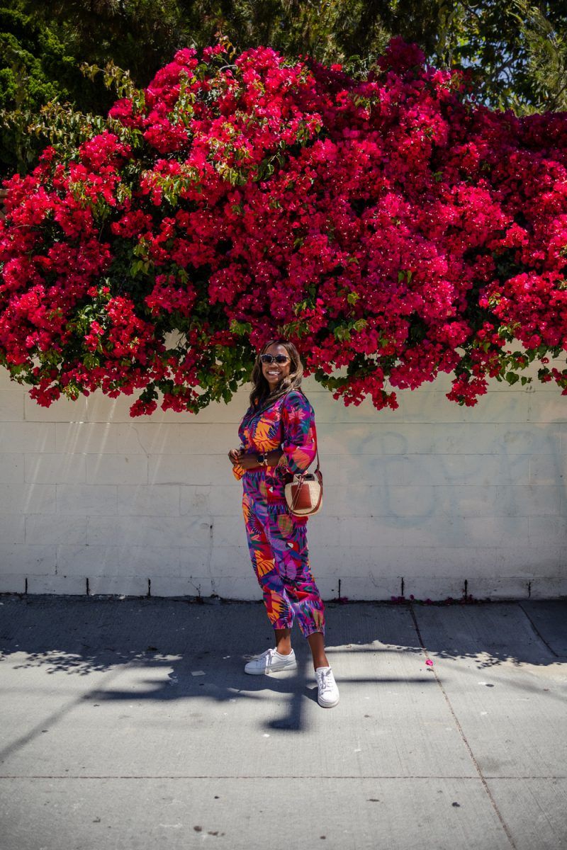 Farm Rio Macaw Twill Jumpsuits | Summer Jumpsuit by popular LA fashion blogger, Alicia Tenise: image of a woman wearing a summer jumpsuit with a pair of white sneakers and standing under a pink floral plant.