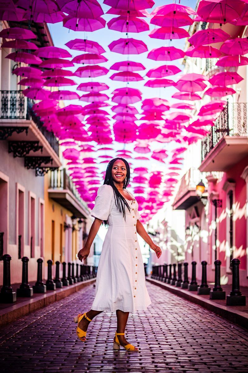Tom's Travel Photography Tips: How to Nail the Perfect Shot While Traveling by popular travel blogger, Alicia Tenise: image of a woman walking under pink umbrellas that are hanging above a cobblestone street.