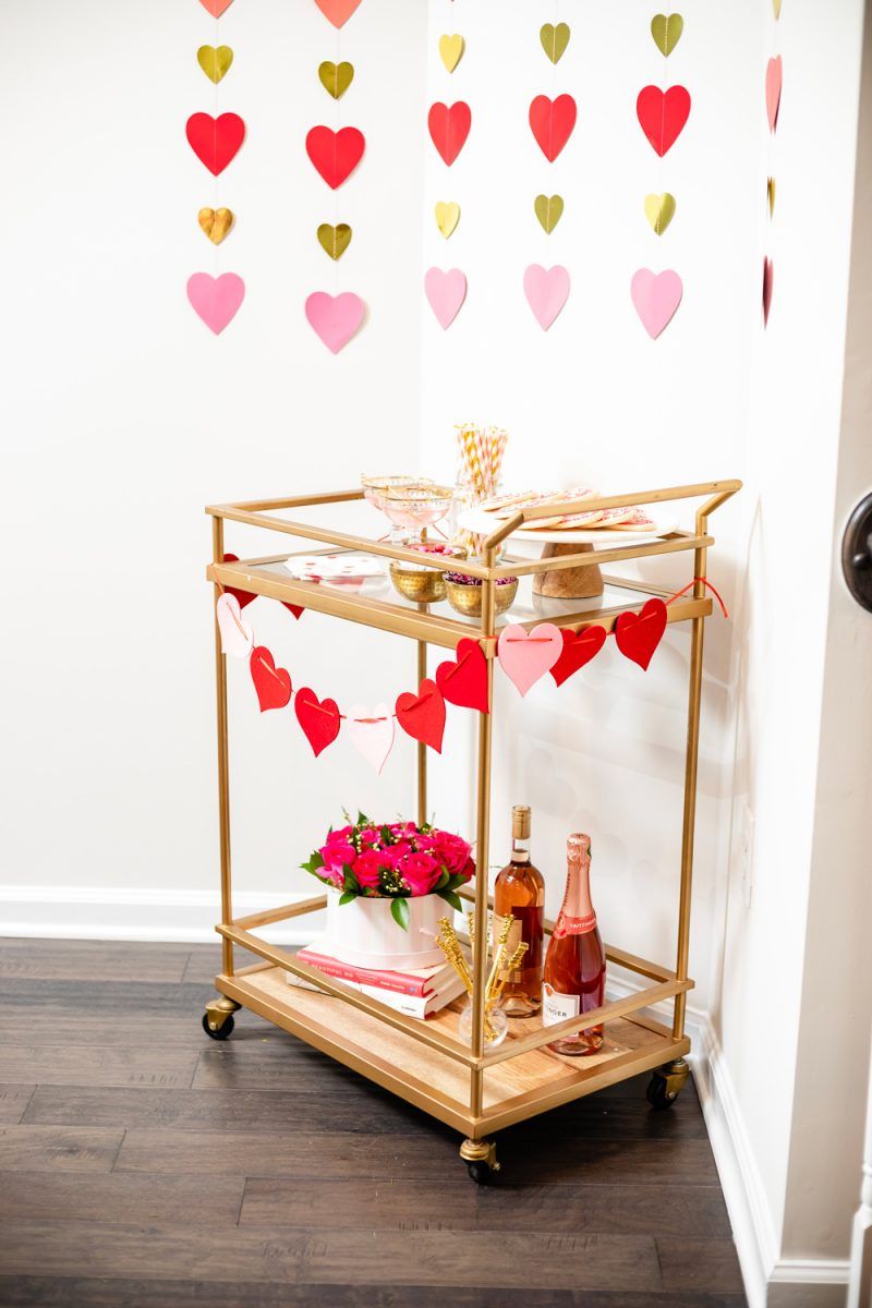 How to Style a Galentine's Day Bar Cart by popular DC lifestyle blogger, Alicia Tenise: image of a Target Threshold Wood & Brass Finish Bar Cart with fresh pink roses, heart shaped sugar cookies, Amazon ZOOYOO pink/gold Paper Straw, heart napkins, heart garland, and alcoholic beverages on it that's in front of a Target Spritz 6ct DIY Valentine's Paper Heart Streamer backdrop.