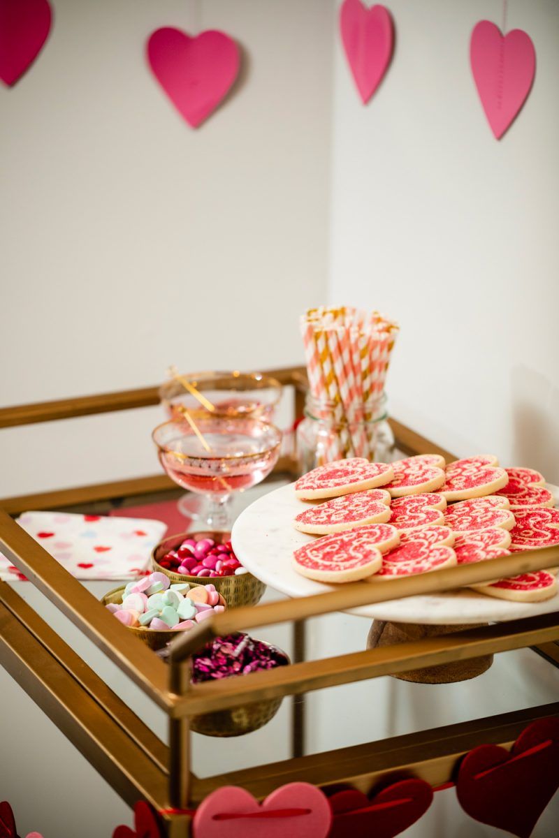 How to Style a Galentine's Day Bar Cart by popular DC lifestyle blogger, Alicia Tenise: image of a Target Threshold Wood & Brass Finish Bar Cart with fresh pink roses, heart shaped sugar cookies, Amazon ZOOYOO pink/gold Paper Straw, heart napkins, glasses, valentine's candy in gold bowls, heart garland, and alcoholic beverages on it that's in front of a Target Spritz 6ct DIY Valentine's Paper Heart Streamer backdrop.