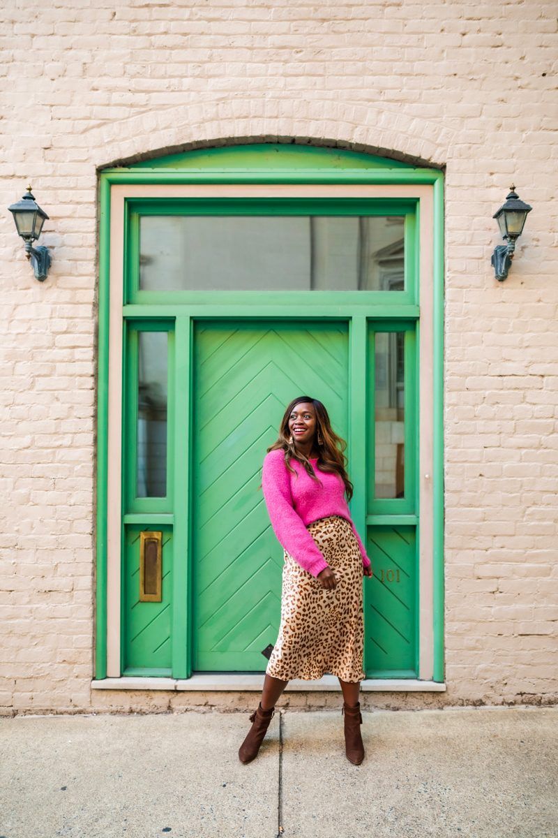 How to Style a Silk Skirt, Free People Cheetah Midi Skirt | Silk Skirt by popular D.C. fashion blogger, Alicia Tenise: image of Alicia Tenise standing in front of a white brick building with a green wooden door and wearing a pink sweater with a Free People Cheetah print silk skirt and brown suede ankle boots.