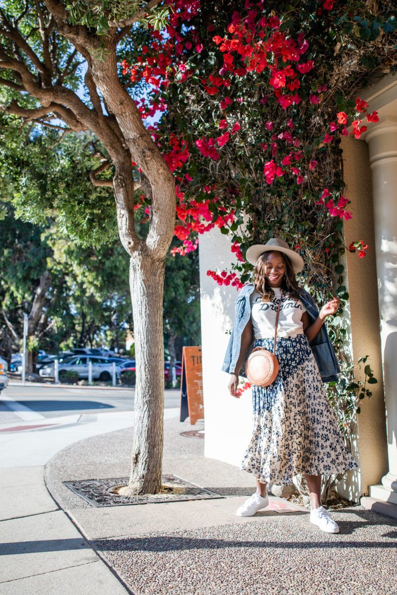 Silk Skirt by popular D.C. fashion blogger, Alicia Tenise: image of Alicia Tenise standing next to a bush with pink flowers and wearing a pleated floral sillk skirt with a felt fedora hat, white sneakers, graphic t-shirt, denim jacket, and circle bag.