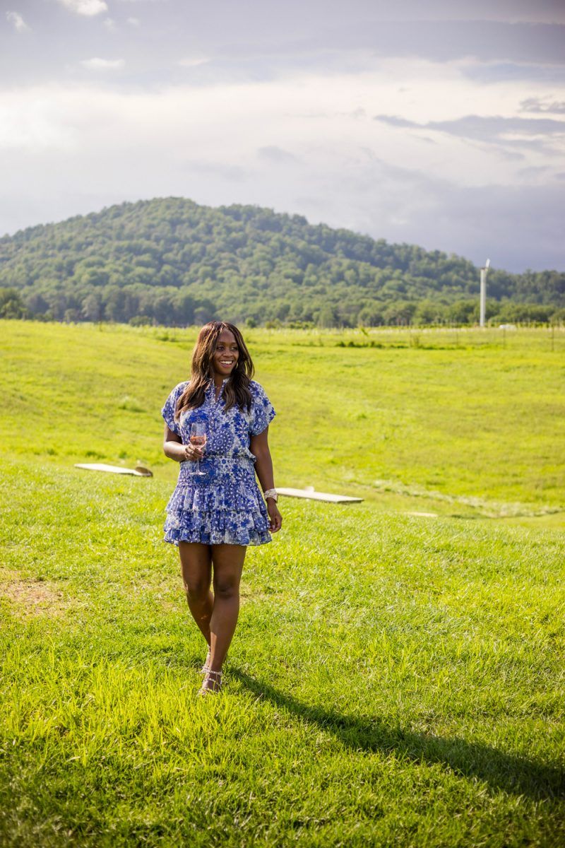 Wine 101 by popular D.C. lifestyle blogger, Alicia Tenise: image of Alicia Tenise standing outside at a winery and wearing a Eloisa Dress MISA Los Angeles brand: MISA Los Angeles, Steve Madden Women's Travel Rock Stud Flat Sandals, GOLD DORADO MIYUKI BEADS APPLE WATCH STRAP, and The Sis Kiss It’s All in a Name™ Personalized Necklace while holding a glass of wine.