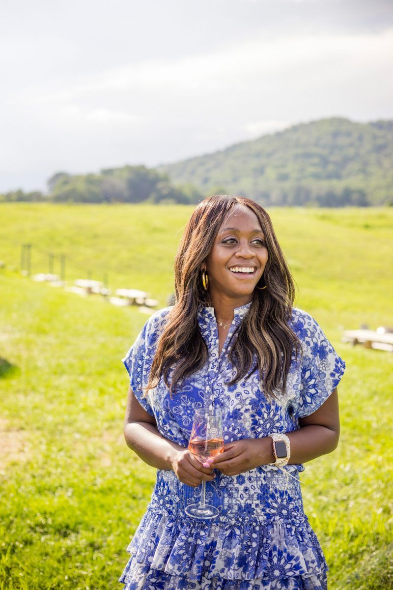 Wine 101 by popular D.C. lifestyle blogger, Alicia Tenise: image of Alicia Tenise standing outside at a winery and wearing a Eloisa Dress MISA Los Angeles brand: MISA Los Angeles, Steve Madden Women's Travel Rock Stud Flat Sandals, GOLD DORADO MIYUKI BEADS APPLE WATCH STRAP, and The Sis Kiss It’s All in a Name™ Personalized Necklace while holding a glass of wine.