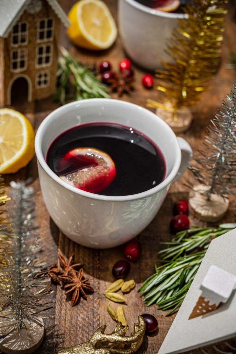 Mulled Wine Recipes by popular D.C. lifestyle blogger, Alicia Tenise: image of mulled wine in a white mug on a table decorated with christmas houses, lemon halves, rosemary springs, and bottle brush christmas trees and whole cranberries.