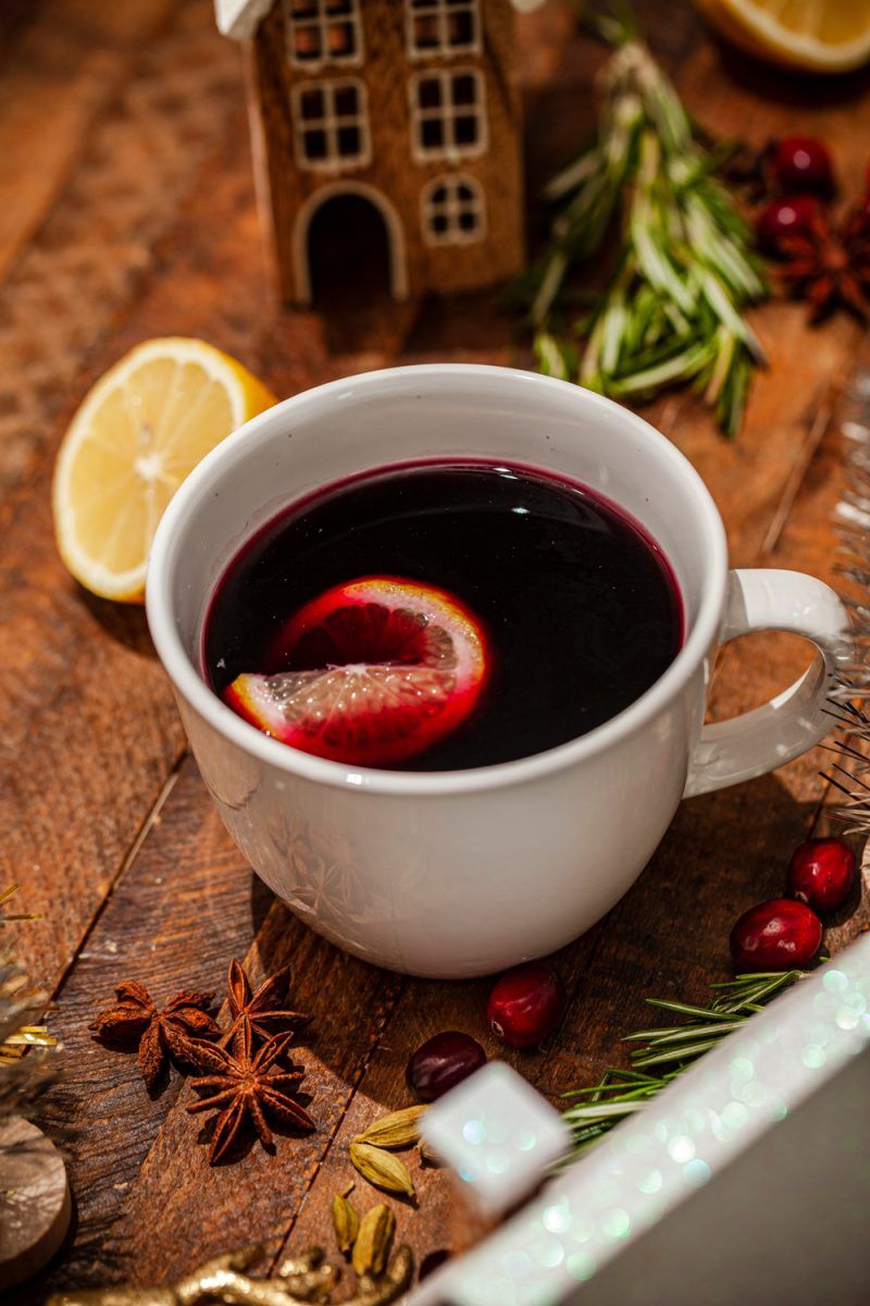 Mulled Wine Recipes by popular D.C. lifestyle blogger, Alicia Tenise: image of mulled wine in a white mug on a table decorated with christmas houses, lemon halves, rosemary springs, and bottle brush christmas trees and whole cranberries.